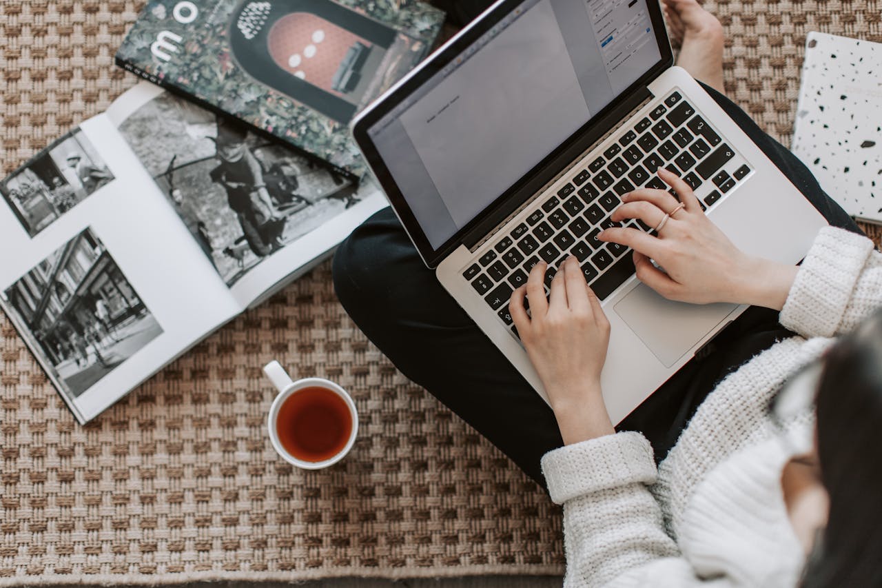 Overhead view of a woman typing on a laptop at home, surrounded by magazines and a cup of tea on the floor.