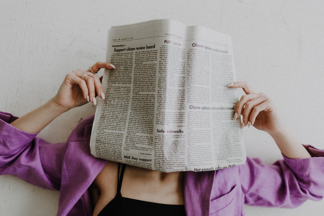 A woman in a violet jacket holds a newspaper, obscuring her face against a light background.